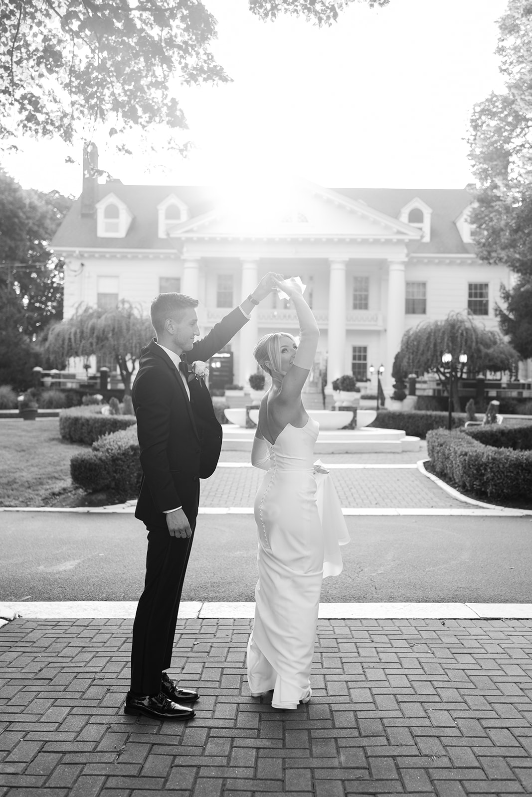 Black and white photo of the groom twirling the bride on a brick pathway, in front of the grand mansion.
